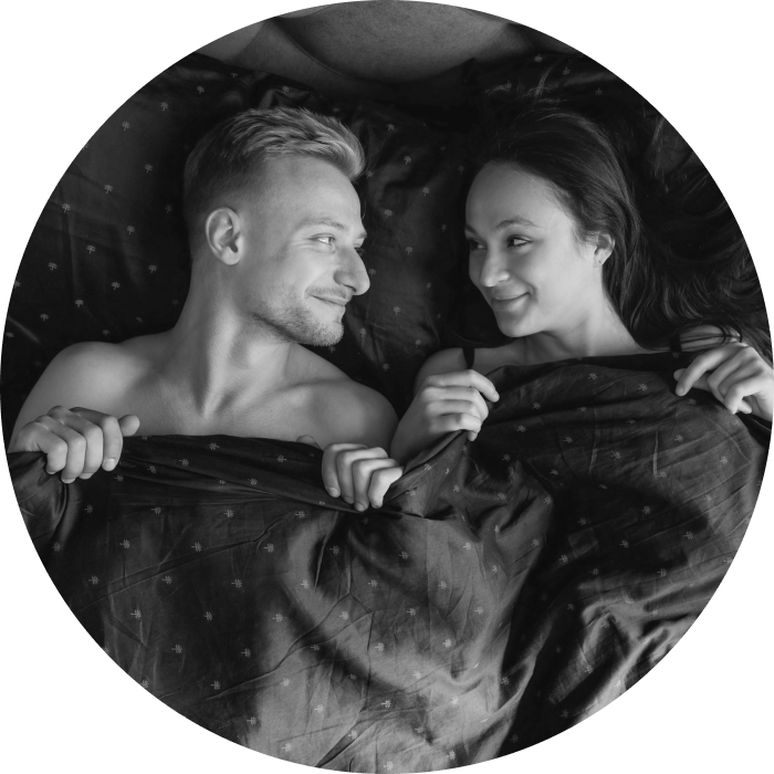 Caucasian couple lying in bed under sheets, sharing a playful, cheeky, secretive smile after intimacy